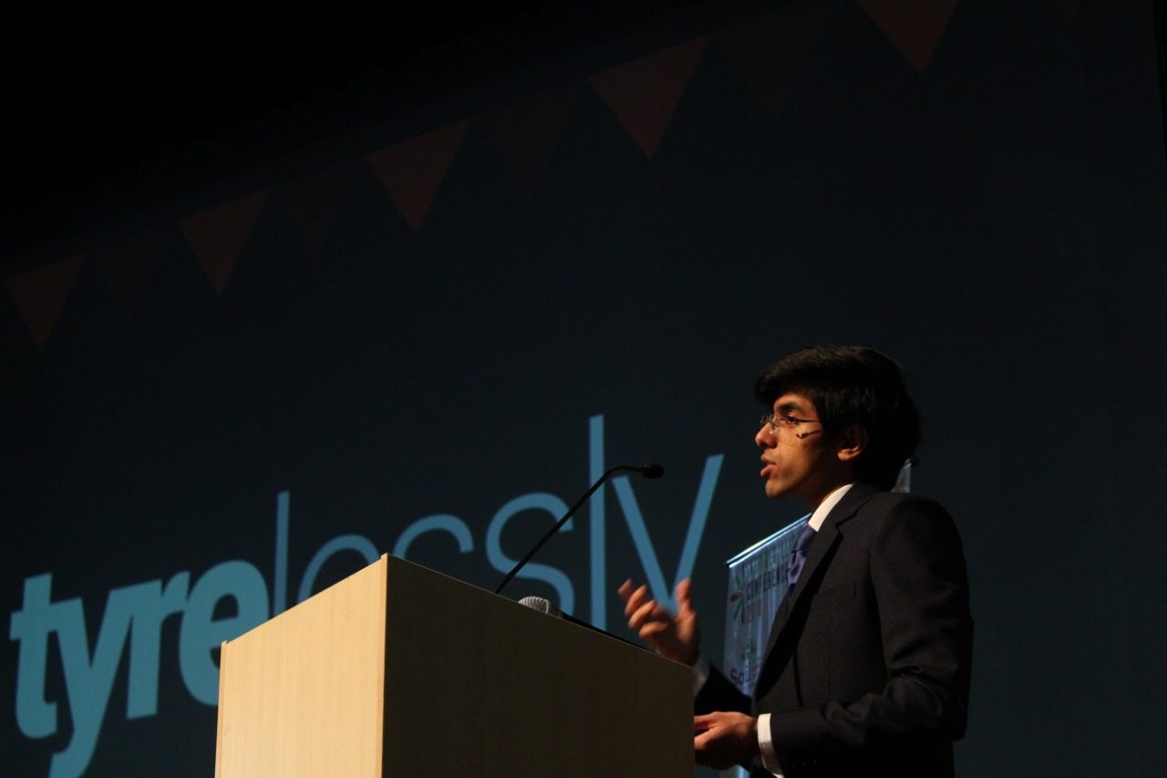 Anubhav Wadhwa speaking at a lectern, with the Tyrelessly logo on the screen behind him